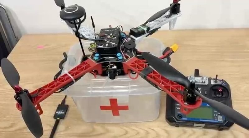 The quadcopter prototype on a bench: red and black airframe over a clear payload box marked with a red cross, with the transmitter beside it.