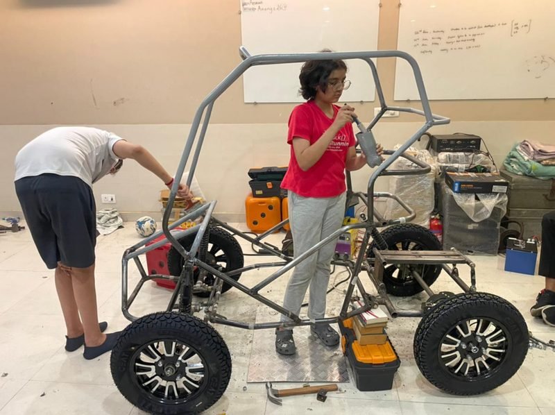 Two students assembling the welded metal roll-cage chassis on four wheels, with tools and a generator on the floor around them.