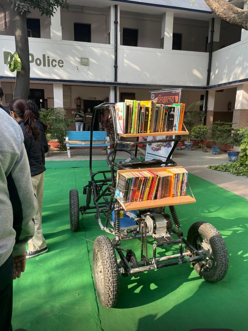 The finished electric vehicle parked on green turf outdoors, fitted with tiered bookshelves full of books in place of a passenger seat.