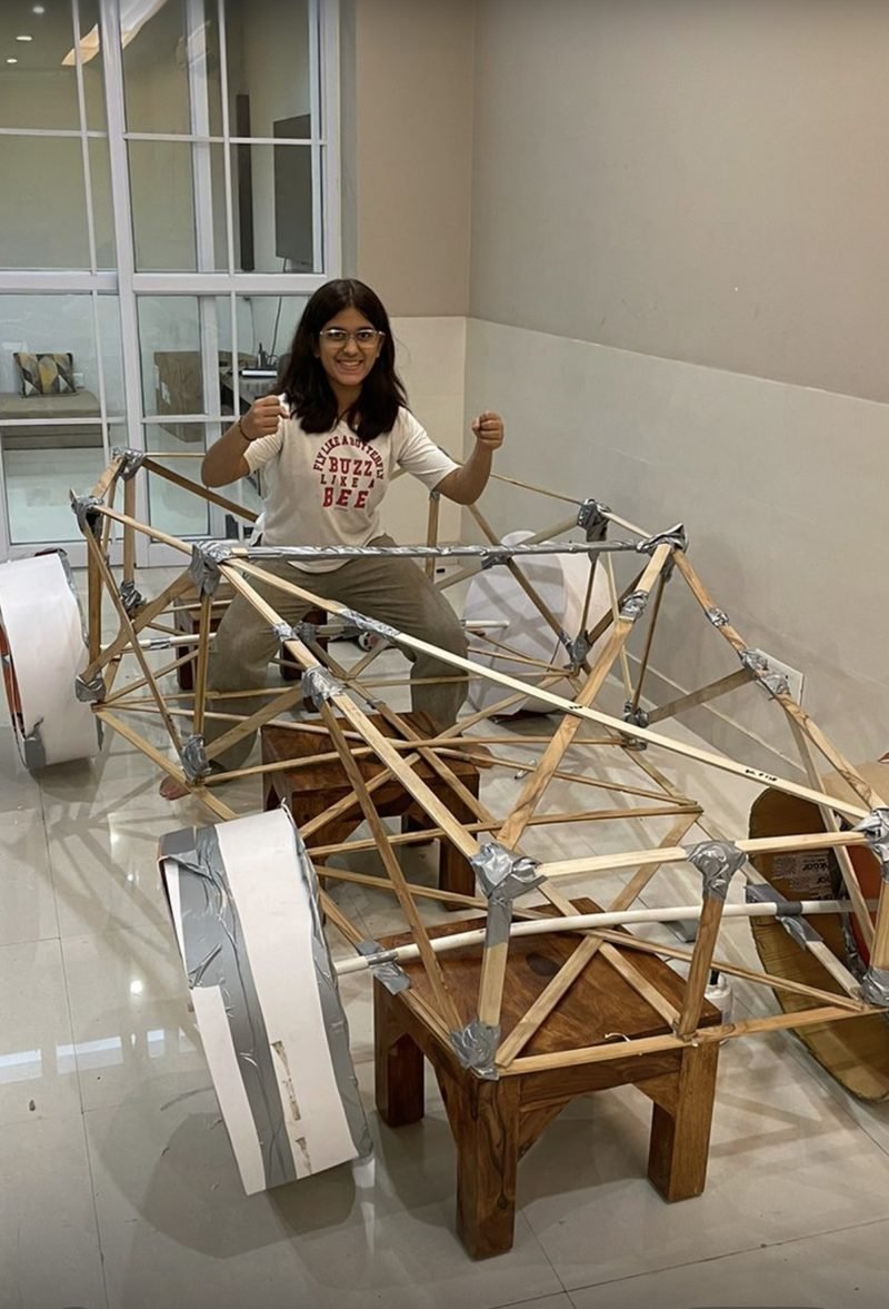 A student standing behind an early full-size vehicle frame built from wooden struts, assembled indoors.